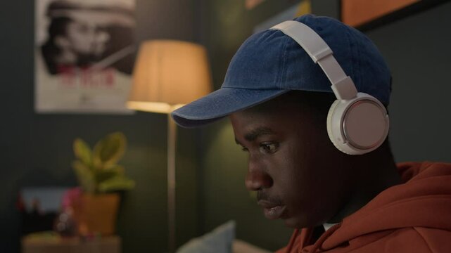 Black boy sitting on sofa in dorm room, listening to music in headphones and using laptop, closeup