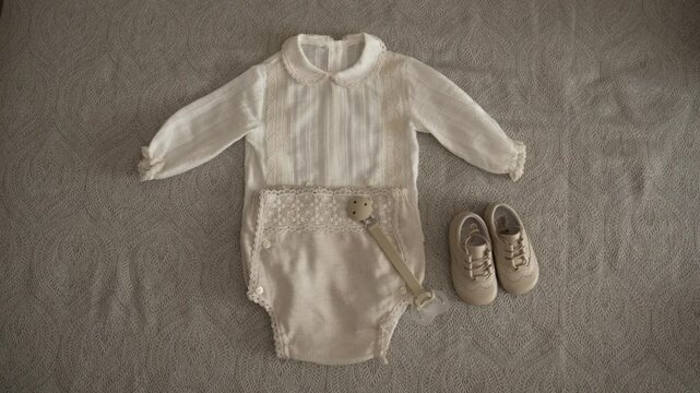 Cream-colored baby outfit with lace details, matching shoes, and a pacifier on fabric