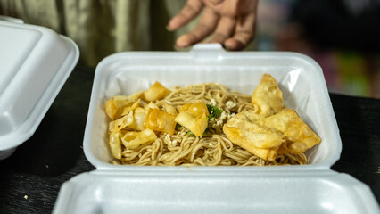 A takeaway box filled with noodles and crispy fried wontons, served in a styrofoam container. The...