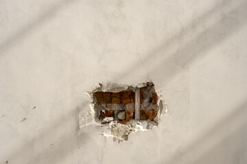 A damaged white plastered wall revealing red bricks and structural reinforcement underneath. Sunlight casts diagonal shadows, adding depth and contrast to the texture of the exposed materials.