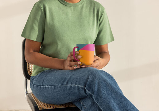 Handmade ceramic mug with handle held by anonyous woman sitting casually on a chair in a minimalist indoor setting. PSD template mockup