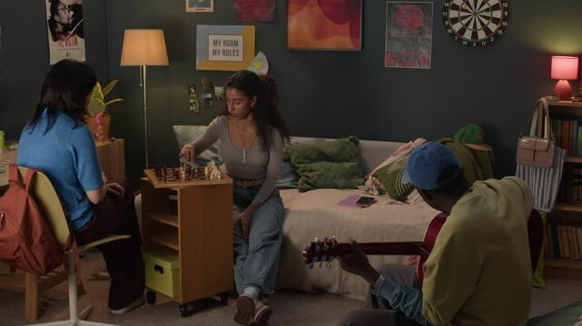 Group of three ethnically diverse friends spending time together in messy dorm room, playing chess and guitar