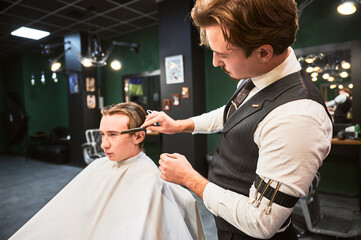 Barber giving haircut to young man in barbershop. Professional barber well-dressed in vest and white shirt with rolled-up sleeves, concentrating on cutting the hair, using comb and scissors.