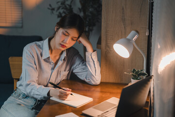 Tired asian businesswoman working late writing on notebook at home office desk at night