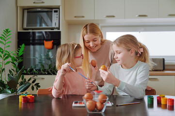 Mother guiding two daughters in meticulously decorating Easter eggs using handmade brushes and...