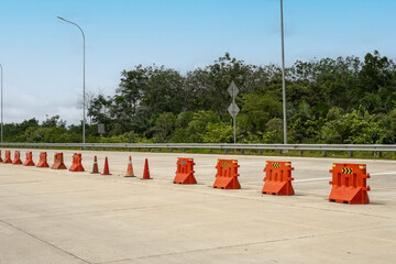 Highway with orange traffic barriers and cones blocking a lane, surrounded by lush green trees. Road safety and construction zone concept under a bright sky.