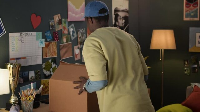 Young Black college student holding box, moving in modern students dormitory room