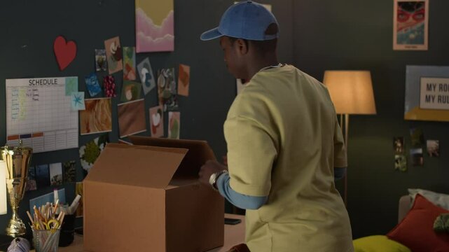 Young African American guy moving in dorm room, standing at table and unpacking cardboard box
