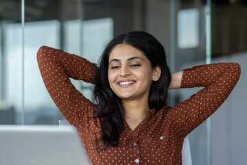 Successful businesswoman resting at workplace inside office. Woman has her hands behind her head and looks at laptop screen, relaxes while sitting at desk.