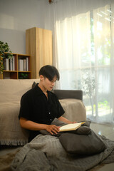 Calm asian man sitting on floor in bright living room and reading book