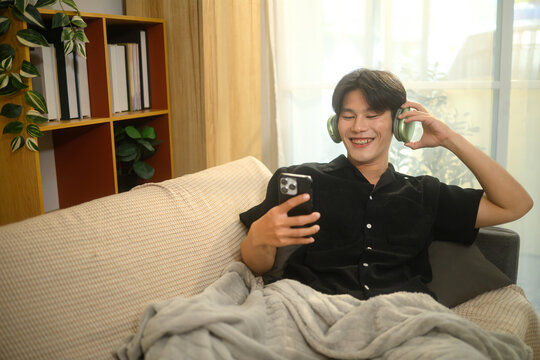 Cheerful young asian man wearing wireless headphones and watching something on smartphone