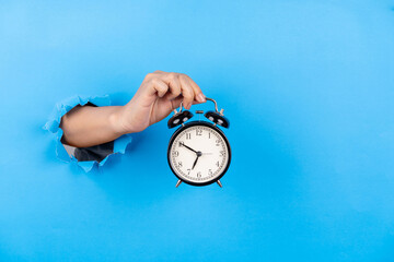 Female hand holding alarm clock through a blue paper background