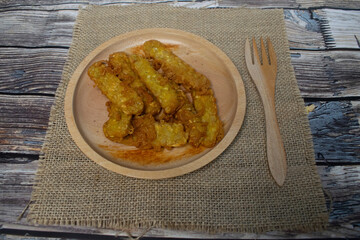 A wooden plate containing flour fried tempeh is served on a burlap cloth on a wooden background