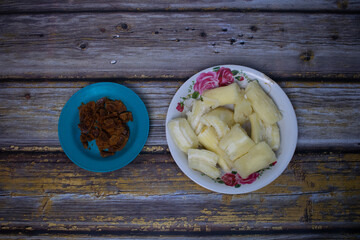 A plate of boiled cassava cut into pieces is served on a floral patterned plate, accompanied by brown sugar
