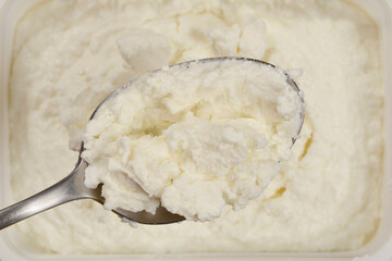 A spoonful of creamy white cottage cheese is lifted from a rectangular containe showcasing its...