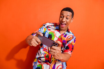 Joyful young man in vibrant shirt holding phone against orange backdrop, embodying cheerful energy...