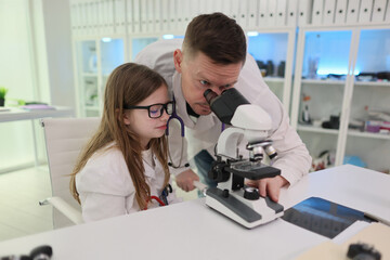 In a lab, a young girl observes through a microscope, guided by an adult, experiencing handson scientific discovery