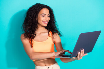 Smiling woman holding laptop against turquoise background, showcasing a casual style and trendy...