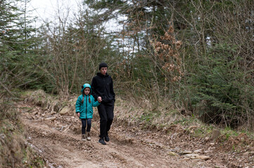 Obraz premium Exploring a serene forest trail, a child in a teal jacket walks hand in hand with an adult, both surrounded by lush greenery on a cool day in late autumn