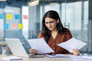 A frustrated young woman with glasses looks over paperwork at her desk in a modern office,...