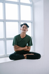 Fototapeta premium Meditative young man sitting near window in relaxed pose indoor studio