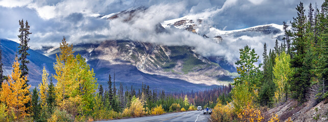 Scenic mountain road in the Canadian Rockies surrounded by autumn trees and misty peaks. A breathtaking view of nature’s beauty with dramatic clouds over snow-capped mountains.