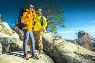 Two hikers, a man and a woman, standing on rocky mountain terrain wearing bright outdoor clothing,...