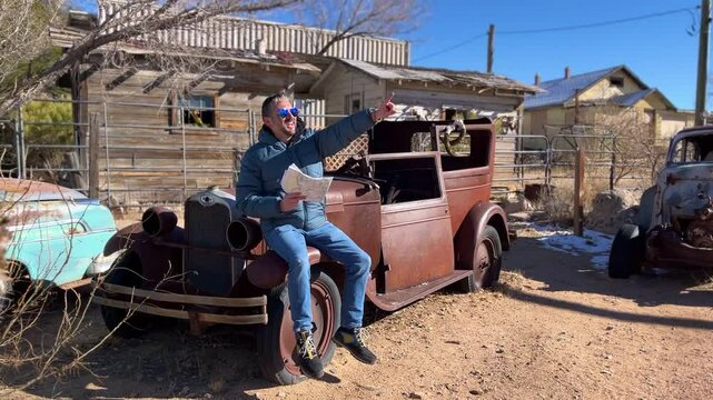 Traveler navigating route through abandoned desert landscape, studying map while perched on weathered vintage automobile in historic Route 66 ghost town setting