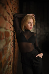 Fototapeta premium A beautiful blonde woman in a black bodysuit and pants posing near a brick wall.