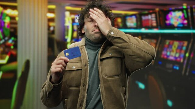 Man indoors holding credit card in casino with slot machines in view, expressing worry suggesting financial stress or gambling loss.