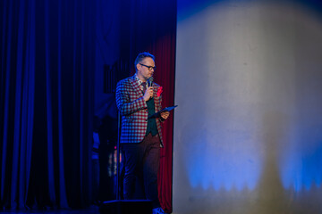A male presenter in a checkered suit holds a microphone and clipboard on stage under blue lighting, delivering a speech or performance..
