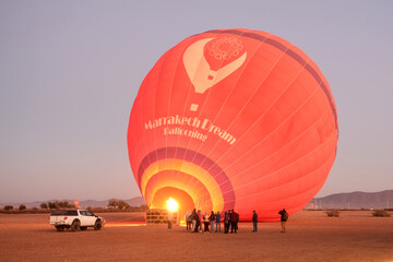 A hot air balloon inflating at sunrise in Marrakech, Morocco, with tourists nearby. The balloon glows from the burner’s flame, preparing for takeoff.