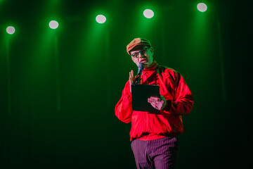 A man in a red jacket and cap speaks into a microphone on stage under green lights, holding a...