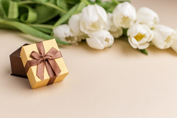 Gift box with brown ribbon beside white tulips on soft surface. Copy space