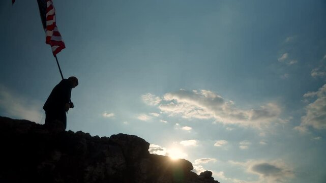Silhouette of young man against the sunlight walking proudly on mountain with holding USA flag with blue sky and sunset background, proud American holding the United states flag celebration concept.
