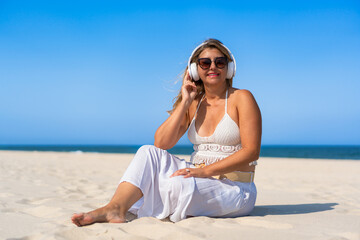 Holidays on beach with music. Beautiful middle aged woman listening to music in headphones sitting on white sandy beach. Relaxing in summer