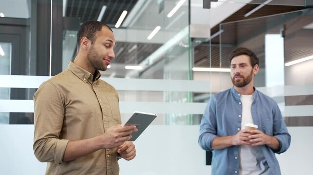 Two coworkers greet each other with a handshake in a modern business office. Happy smiling business colleagues have friendly conversation during break. Workers enjoying casual discussion with coffee