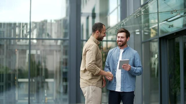 Two businessmen shaking hands outside modern office building, symbolizing successful business partnership and teamwork. Colleagues co-workers portray professional cooperation and friendly interaction