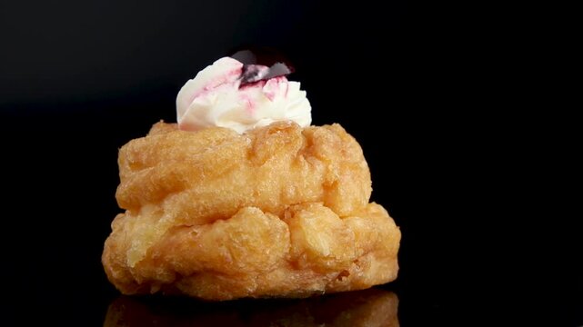 A delicious Italian Bigne alla Crema pastry spins on a black background. This choux pastry filled with a creamy mix of whipped cream and ricotta, topped with a cherry, looks irresistible.
