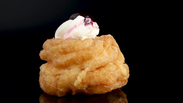 A delicious Italian Bigne alla Crema pastry spins on a black background. This choux pastry filled with a creamy mix of whipped cream and ricotta, topped with a cherry, looks irresistible.