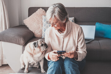 Smiling senior woman sitting on sofa at home with her old  pug dog having relaxing moments. Therapy...