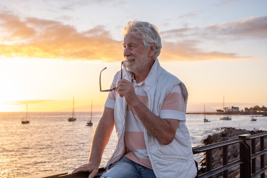 Smiling bearded senior man standing at sunset light on sea shore, carefree senior male enjoying vacation, free time and retirement