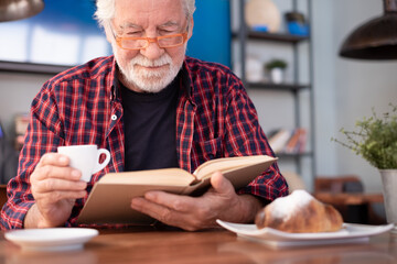 Smiling relaxed senior bearded man sitting at cafe table reading a book enjoying breakfast with coffee and croissant. Elderly caucasian male in eyeglasses and checkered shirt