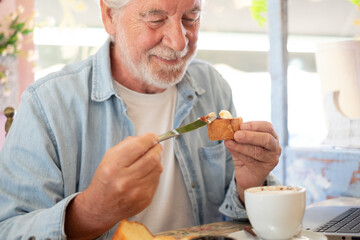 Portrait of smiling bearded senior man having breakfast in coffee shop with sweet food and cappuccino while works on laptop. Elderly man people enjoying free lifestyle doing job from remote