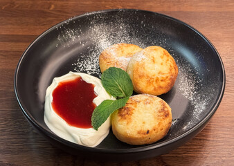 Close-up photo delicious healthy breakfast of cheesecakes with sour cream, strawberry jam and mint leaves lying in a black plate on a wooden table