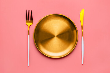 Golden plate with matching fork and knife arranged on a pink background, creating a stylish and...