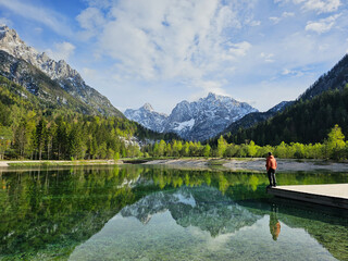 Hiker standing on wooden pier overlooking jasna lake, reflecting snow-dusted julian alps peaks in slovenia's triglav national park during tranquil spring sunrise