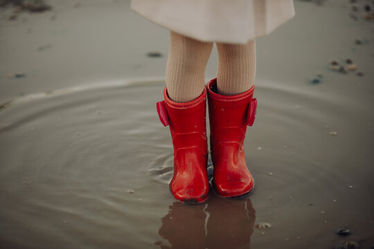 a child stands in a puddle in beautiful red rubber boots. a walk in rainy weather, slush and mud on the road, spring and autumn. children's favorite pastime
