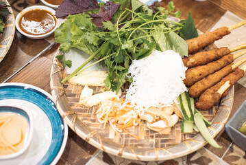 Authentic charcoal grilled lemongrass pork skewers “Nem Lui Hue” on bamboo mat with Vietnamese rice noodle, cucumber, ginger, carrot slices, pineapple, lettuce, cilantro and herbs, dipping sauce