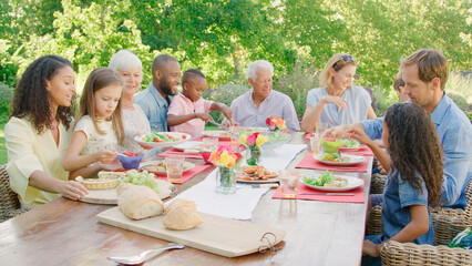 Multi-Generation Family With Friends Sitting Around Outdoor Table In Garden Enjoying Meal Together